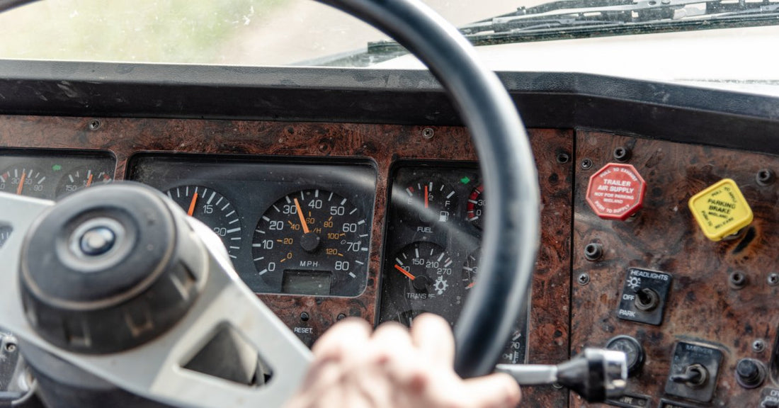 A semitruck dashboard with different meters, buttons, and a steering wheel. There is a hand clutching the steering wheel.