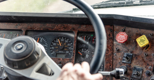 A semitruck dashboard with different meters, buttons, and a steering wheel. There is a hand clutching the steering wheel.