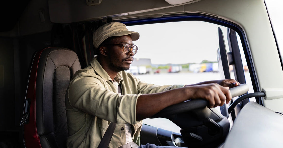 A man operating a semi-truck with both hands on the steering wheel. He is wearing a beige hat and a long-sleeved shirt.