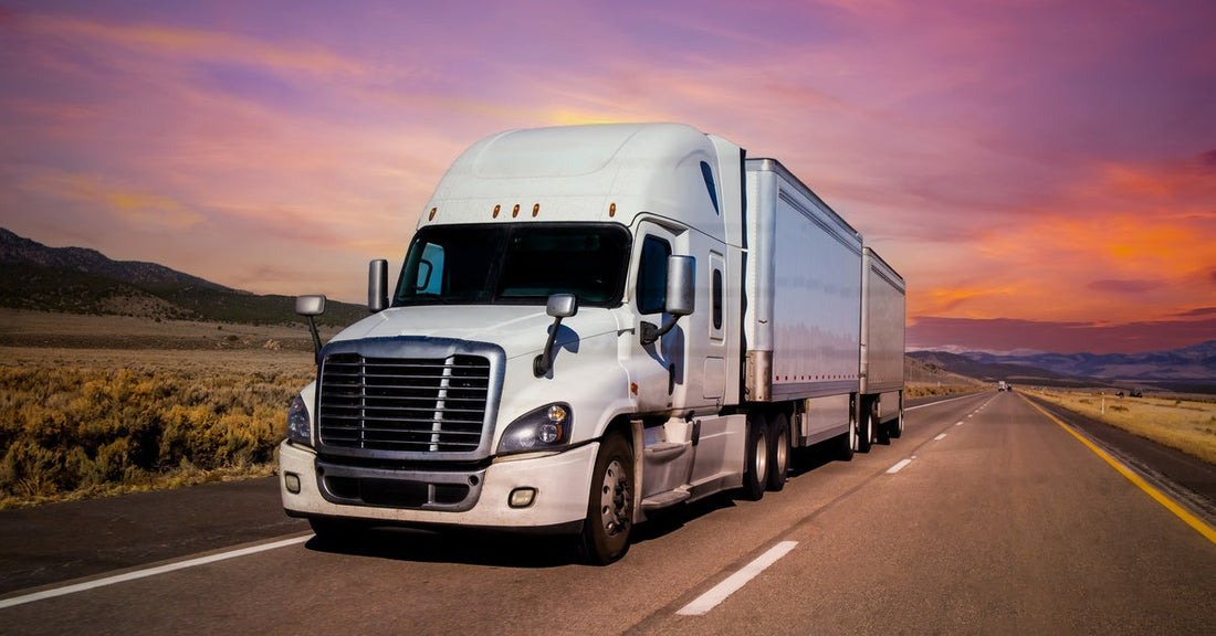 A white semi-truck driving down a two-lane highway. The sky is full of pink, purple, and orange clouds.
