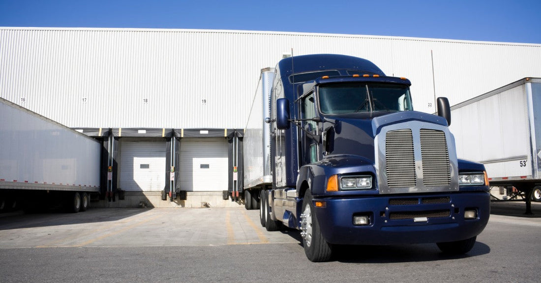 A dark blue semi-truck parked at a docking station outside of a warehouse. There are other semi-trucks parked nearby.