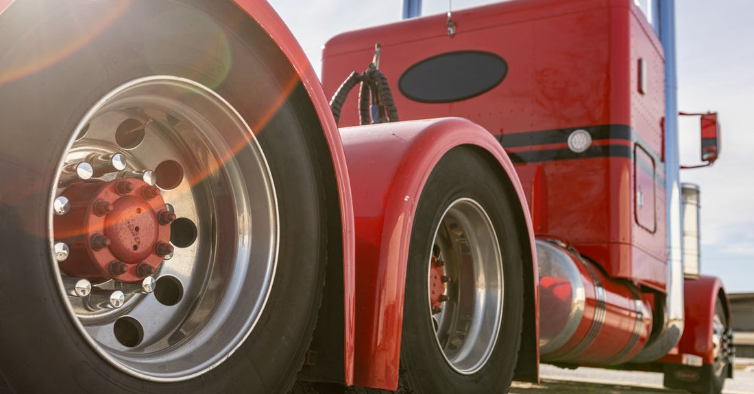 The rear side of a classic red semitruck parked in a lot. The sunlight bounces off of the truck’s chrome rims.