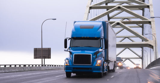 A blue semi-truck driving underneath an arched bridge with its headlights on. Behind the truck, there are other cars driving.