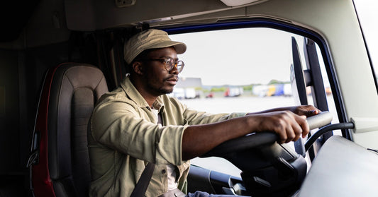 A man operating a semi-truck with both hands on the steering wheel. He is wearing a beige hat and a long-sleeved shirt.