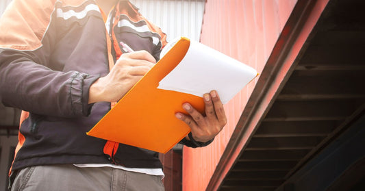 A person holds an orange clipboard and writes on a sheet of paper. A semitruck is parked beside them.