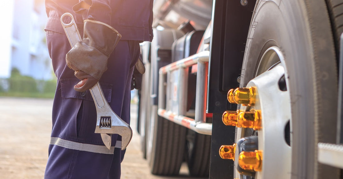 A mechanic holds a wrench and stands in front of a large tire on a semi-truck. The truck is parked on a paved area.