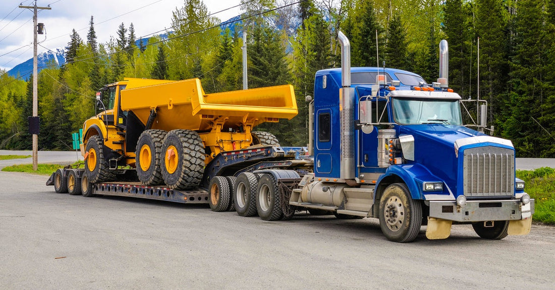 A blue tractor unit drives down a road with trees and is hauling a yellow dump truck. The machinery is fixed to the trailer.