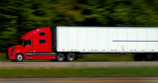 A red-cabbed semi-truck is driving down a tree-lined highway. It's pulling a white trailer with a blue stripe.