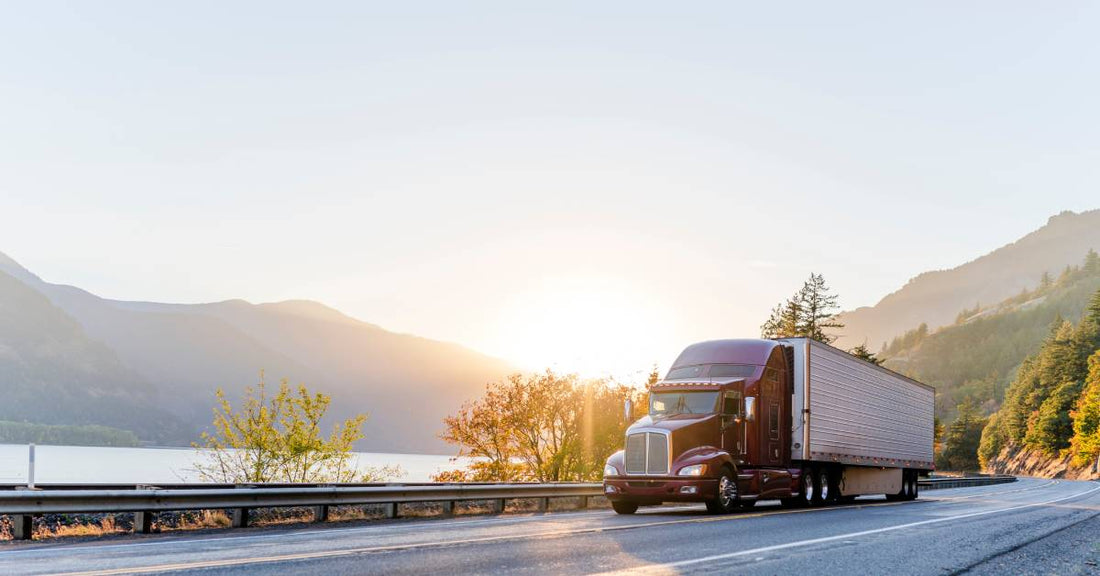 A semi-truck with a dark red cab and white trailer travels down a two-lane road surrounded by a lake and mountains.