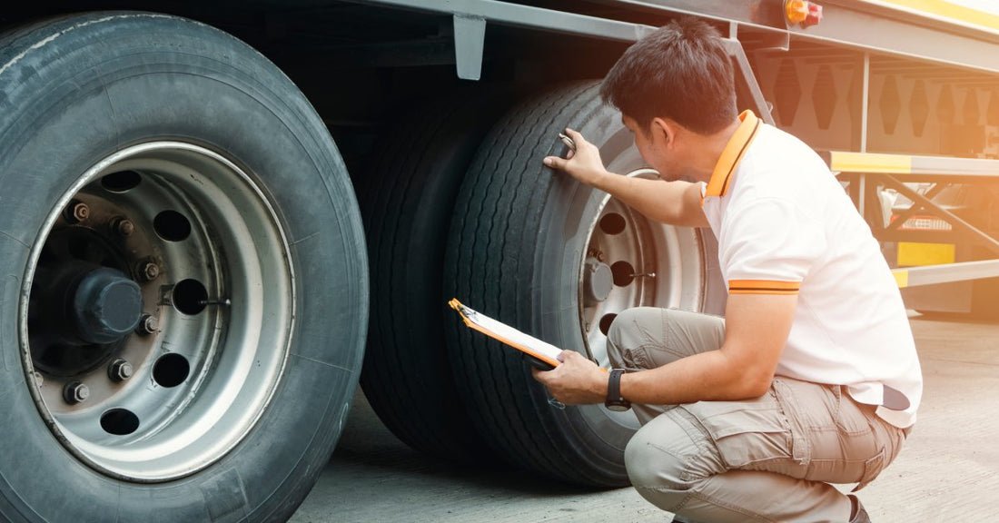 A mechanic kneeling to inspect the tires on a semi-truck. He holds a clipboard in one hand and touches a tire with the other.