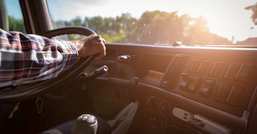 A truck driver in a plaid shirt behind the steering wheel and cabin dashboard of his commercial-grade semi-truck.